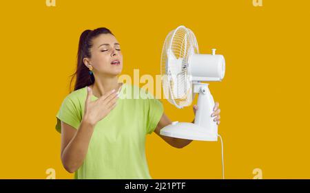 Erschöpfte junge Dame, die wegen der Sommerhitze Kopfschmerzen hat, die Fan in der Hand hält, ist von der Hitze erschöpft, berührt ihre Stirn, gelbes Studio Stockfoto