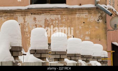 Hohe, wunderschöne Schneesäulen auf Ziegelsteinsäulen. Eine gerade Reihe kleiner Schneeverwehungen vor dem Hintergrund einer schäbigen Wand. Urbane Winterlandschaft. Wolkiger Wintertag, weiches Licht. Stockfoto