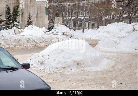 Lockere große Schneeverwehung an der Straße vor dem Hintergrund einer Stadtstraße. Auf der Straße liegt schmutziger Schnee in hohen Haufen. Urbane Winterlandschaft. Wolkiger Wintertag, weiches Licht. Stockfoto