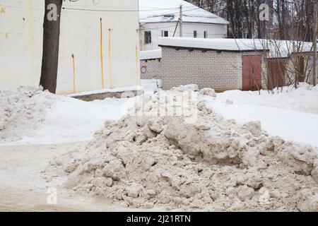 Eine große Schneeverwehung an der Straße vor dem Hintergrund einer Stadtstraße. Auf der Straße liegt schmutziger Schnee in hohen Haufen. Urbane Winterlandschaft. Wolkiger Wintertag, weiches Licht. Stockfoto