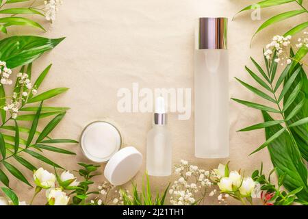 Nachbau von Beauty-Produkten. Kosmetikflaschen aus Glas, offenes Cremeglas, tropische Pflanzen und Blumen auf Sandgrund. Weiches Bild und weicher Fokus. O Stockfoto