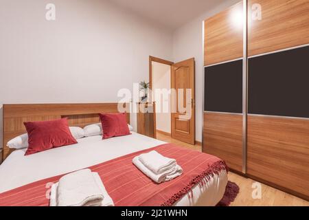 Schlafzimmer mit Kingsize-Bett, roten Kissen, passender Decke, weißer Bettwäsche und Kleiderschrank mit Schiebetüren aus Holz Stockfoto