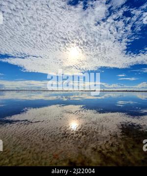 Ein atemberaubender Blick auf die helle Sonne, die hinter den Altocumuluswolken im blauen Himmel verborgen ist, der sich im Meer widerspiegelt Stockfoto