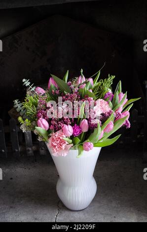 Ein Bouquet aus rosa Tulpen, Chrysanthemen und Heidekraut in einer weißen Vase in einem rustikalen Kamin. Stockfoto