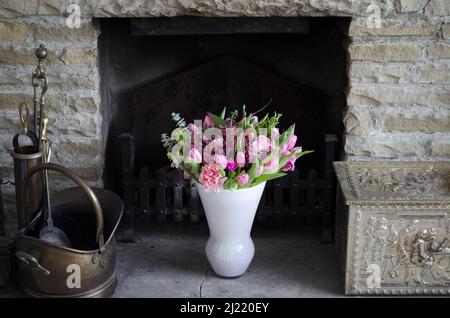 Ein Bouquet aus rosa Tulpen, Chrysanthemen und Heidekraut in einer weißen Vase in einem rustikalen Kamin. Stockfoto