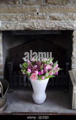 Ein Bouquet aus rosa Tulpen, Chrysanthemen und Heidekraut in einer weißen Vase in einem rustikalen Kamin. Stockfoto