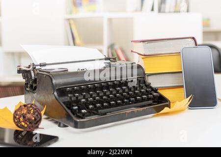 Arbeitsplatz eines Schriftstellers oder Korrespondenten: Eine alte Schreibmaschine, ein Mobiltelefon, Bücher und zwei gelbe, trockene Ahornblätter Stockfoto