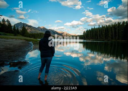 Frau, die im flachen Wasser des Johnson Lake im Banff National Park steht und Berge und ländliche Szenen betrachtet. Stockfoto