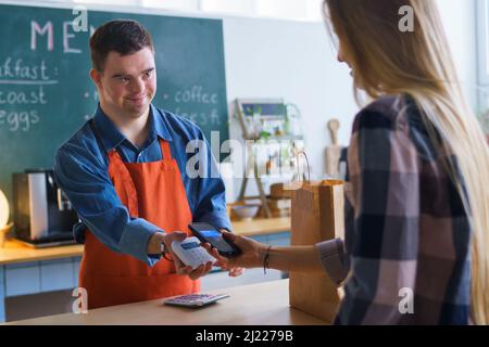 Fröhlicher junger Down-Syndrom-Kellner, der kontaktlose Smartphone-Zahlung vom Kunden im Take-Away-Restaurant nimmt. Stockfoto