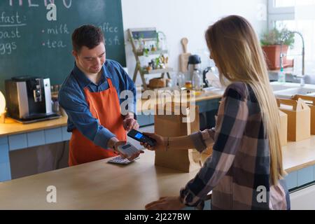 Fröhlicher junger Down-Syndrom-Kellner, der kontaktlose Smartphone-Zahlung vom Kunden im Take-Away-Restaurant nimmt. Stockfoto