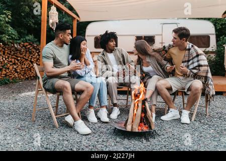 Verschiedene junge Freunde sitzen am Lagerfeuer und erzählen während des Campingurlaubs neben dem Wohnmobil unheimliche Geschichten Stockfoto