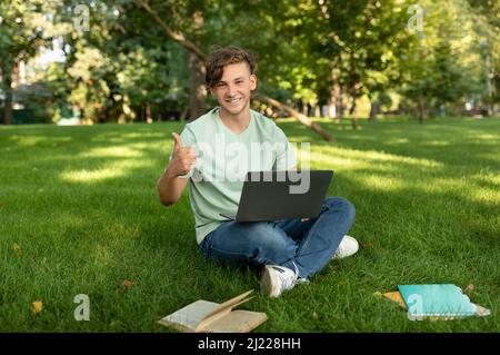 Glücklicher männlicher Student, der wie gestikuliert, das Online-Bildungsprogramm genehmigt, mit einem Laptop im Park sitzt und den Daumen nach oben zeigt Stockfoto