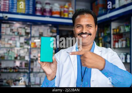 Lächelnder Apotheker in Uniform zeigt grünes Handy, indem er mit dem Finger auf die Apotheke zeigt - Konzept der Online-Buchung, Werbung Stockfoto