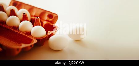 Auf einem weißen Tisch befindet sich ein offenes orangefarbenes Pappkarton mit Hühnereiern in einer weißen Schale, daneben liegen zwei Eier. Gesundes Frühstück. Stockfoto