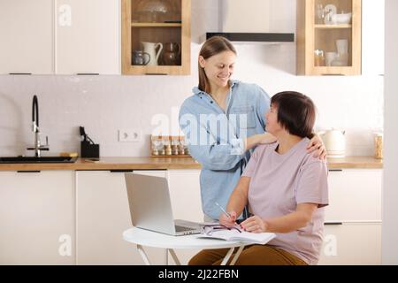 Junge Frau, die ältere Mutter lehrt, das Internet auf dem Laptop zu Hause zu nutzen. Tochter hilft ihrer älteren Mutter, es online mit ihrem persönlichen Konto herauszufinden Stockfoto