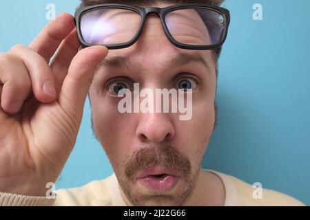 Junger Mann mit witzigem Schnurrbart in großer Brille, der Zweifel hat und mit verwirrem Gesichtsausdruck auf isoliertem blauem Hintergrund Stockfoto