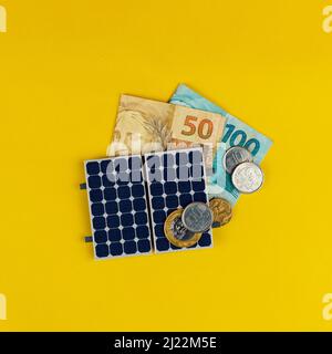 Photovoltaik-Panel , Geld und Steckdraht isoliert auf gelbem Hintergrund. Solarpanel mit variierten Werten des brasilianischen Geldes. Alternativer sauberer Energiebus Stockfoto