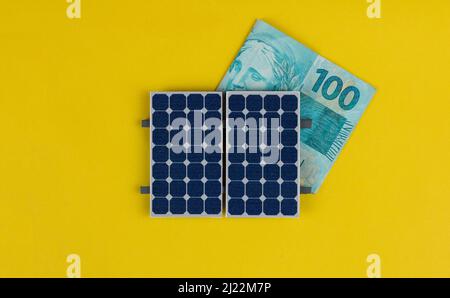 Photovoltaik-Panel , Geld und Steckdraht isoliert auf gelbem Hintergrund. Solarpanel mit variierten Werten des brasilianischen Geldes. Alternativer sauberer Energiebus Stockfoto