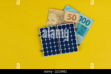 Photovoltaik-Panel , Geld und Steckdraht isoliert auf gelbem Hintergrund. Solarpanel mit variierten Werten des brasilianischen Geldes. Alternativer sauberer Energiebus Stockfoto