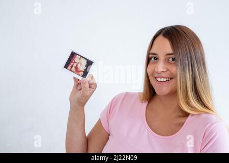 Porträt Schwangeren jungen kaukasischen Frau drinnen lächelnd Blick auf die Kamera zeigt Ultraschall-Druck des Babys auf weißem Hintergrund Stockfoto