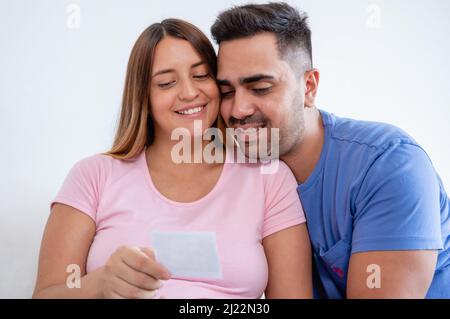 Glücklich schwanger kaukasischen jungen Mann und Frau sitzen auf dem Bett im Schlafzimmer und beobachten Ultraschall-Foto mit weißer Wand auf dem Hintergrund. Stockfoto