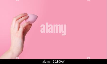 Banner mit Hand hält Make-up Schwamm oder Mixer auf rosa Hintergrund. Schönheitskonzept. Auftragen von Fundament, Concealer, Balsam. Gesichtskonturierung. Speicherplatz kopieren. Hochwertige Fotos Stockfoto