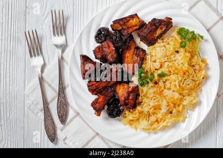 Geschmortes Sauerkraut mit Schweinefleisch und Pflaumen auf weißem Teller auf Holztisch, horizontale Ansicht von oben, flaches Lay, Nahaufnahme Stockfoto