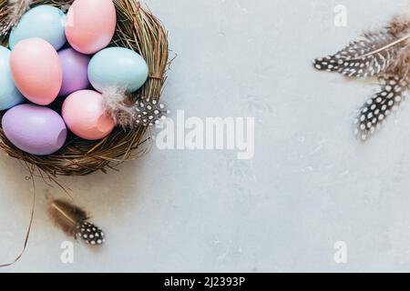 Happy Easter Konzept. Vorbereitung auf den Urlaub. Farbenfrohe, pastellfarbene Ostereier im Nest mit Federn auf betonsteingrauem Hintergrund. Einfacher Minimalismus flach Lay Draufsicht Kopieplatz Stockfoto