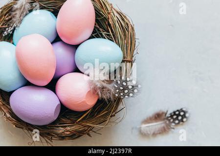 Happy Easter Konzept. Vorbereitung auf den Urlaub. Farbenfrohe, pastellfarbene Ostereier im Nest mit Federn auf betonsteingrauem Hintergrund. Einfacher Minimalismus flach Lay Draufsicht Kopieplatz Stockfoto
