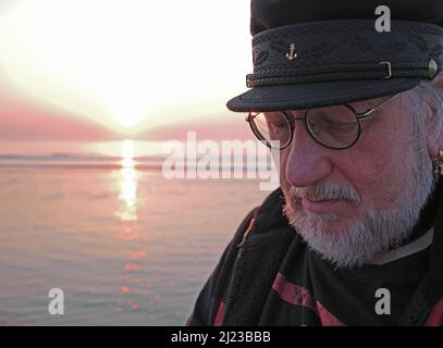 Der alte Mann und das Meer bei Sonnenuntergang. Ein älterer Mann schaut nachdenklich nach unten. Der Mann hat einen weißen Bart und eine Matrosenmütze auf dem Kopf Stockfoto