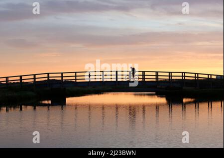 Farbfoto des Radfahrers, der bei Sonnenaufgang über der Brücke über Afon Crigyll in Rhoseigr an der Westküste der Isle of Anglesey, North Wales U, übergeht Stockfoto