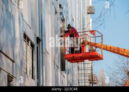 Dnepropetrovsk, Ukraine - 02.09.2022: Baumeister in einem Krankorb repariert die Beschichtung einer Metallstruktur an der Wand der Fassade eines Gebäudes. Stockfoto