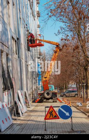 Dnepropetrovsk, Ukraine - 02.09.2022: Baumeister in einem Krankorb repariert die Beschichtung einer Metallstruktur an der Wand der Fassade eines Gebäudes. Stockfoto