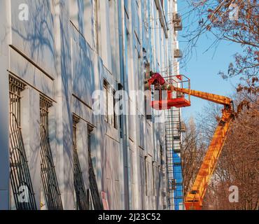 Dnepropetrovsk, Ukraine - 02.09.2022: Baumeister in einem Krankorb repariert die Beschichtung einer Metallstruktur an der Wand der Fassade eines Gebäudes. Stockfoto