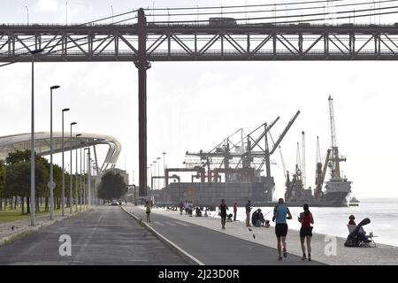 Menschen an der Promenade und Fischer am Fluss Tejo in der Nähe der Brücke 25 de Abril mit Industriehafen im Hintergrund, Lissabon Portugal. Stockfoto