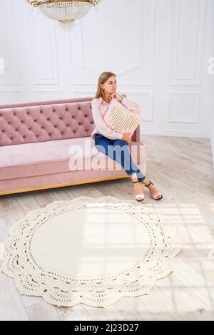 Eine junge Frau sitzt auf einem rosa Sofa und hält ein selbstgestricktes Kissen in den Händen. Nadelarbeit Stockfoto