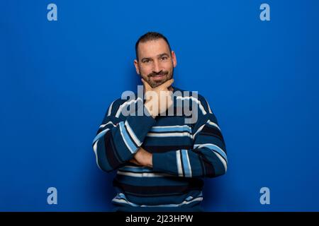 Hübscher bärtiger Mann mit gestreiftem Pullover auf blauem Hintergrund, der mit gekreuzten Armen und erhobener Hand bis zum Kinn die Kamera lächelnd anschaut Stockfoto