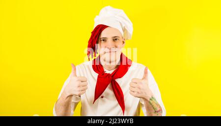 Junger Mann, der als Koch gekleidet war und auf gelbem Hintergrund eine Geste wie ein Gestus zeigte. Männlicher Koch zeigt Daumen nach oben im Studio Stockfoto