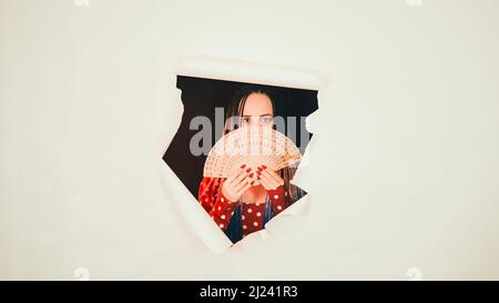 Junge Frau in rotem Kleid mit Fächer in Loch von weißem Hintergrund im Studio. Hübsche Dame, die sich hinter einem Ventilator versteckt und die Kamera anschaut Stockfoto