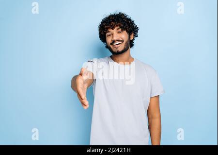 Ein junger Erwachsener, positiv gelockter indischer oder arabischer Mann, trägt ein legeres T-Shirt, bietet freundlich einen Handschlag als Begrüßung, Zustimmung und Begrüßung an, steht auf einem isolierten blauen Hintergrund und lächelt Stockfoto