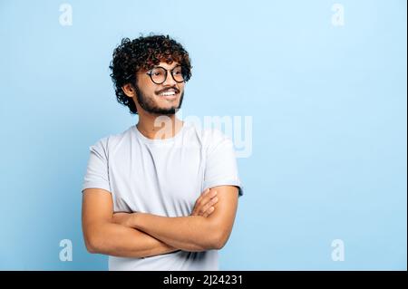 Attraktiver, positiver indischer oder arabischer lockiger Kerl mit Brille, in einem einfachen T-Shirt, Student oder Freiberufler, auf einem isolierten blauen Hintergrund stehend, mit gekreuzten Armen, sieht zur Seite, lächelt Stockfoto