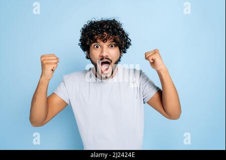 Begeistert staunenden indischen oder arabischen Kerl, in abasischem T-Shirt, freut sich über Erfolg, Sieg, Sieg, Gestikulend mit Fäusten, lächelnd auf die Kamera schauend, auf einem isolierten blauen Hintergrund stehend Stockfoto