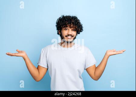 Verwirrt verwirrten indischen oder arabischen lockigen Mann in Freizeitkleidung, zucken Schultern, Blick fragend auf die Kamera, stehend auf isoliertem blauen Hintergrund Stockfoto