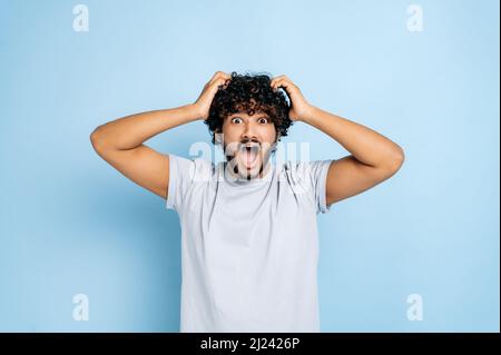 Frustrierter, verängstigt, entmutigter, lockiger indischer oder arabischer Kerl in einem lässigen T-Shirt, der mit den Händen am Kopf hält, verwirrt mit offenem Mund auf die Kamera schaut und über einem isolierten blauen Hintergrund steht Stockfoto