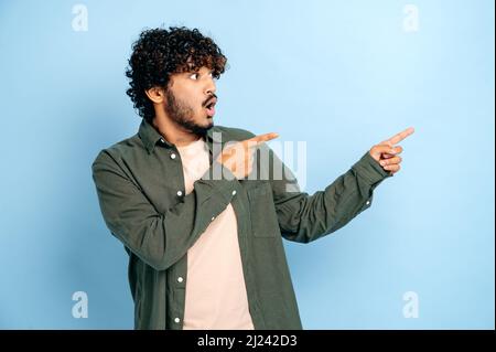 Schockiert überraschte indische oder arabische Lockenhaarige in legerer Kleidung, zeigt die Finger zur Seite auf einen nachgemoßelten leeren Raum, steht auf isoliertem blauem Hintergrund, schaut erstaunt zur Seite Stockfoto