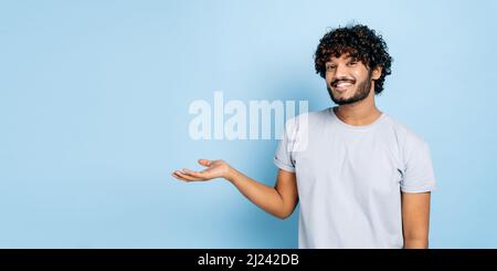 Panoramafoto von freundlichem inder oder araber im lässigen T-Shirt, schaut auf die Kamera und zeigt die Hand zur Seite, auf leeren Raum, steht auf isoliertem blauem Hintergrund und lächelt. Modellkonzept Stockfoto