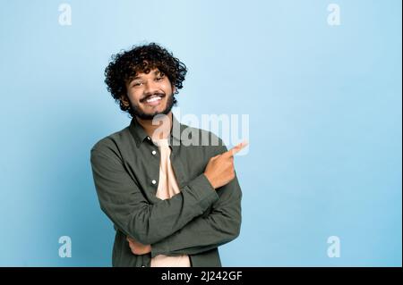 Positiver indischer oder arabischer lockiger Kerl in lässiger Kleidung, zeigt den Finger zur Seite auf den Raum für Ihre Präsentation, steht auf isoliertem blauem Hintergrund, blickt lächelnd auf die Kamera Stockfoto