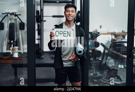 Sie sind jederzeit zwischen 9 und 5 Uhr willkommen. Beschnittenes Porträt eines hübschen jungen Fitnesslehrers, der ein Schild mit der AUFSCHRIFT „OFFEN“ im Stehen hochhält Stockfoto