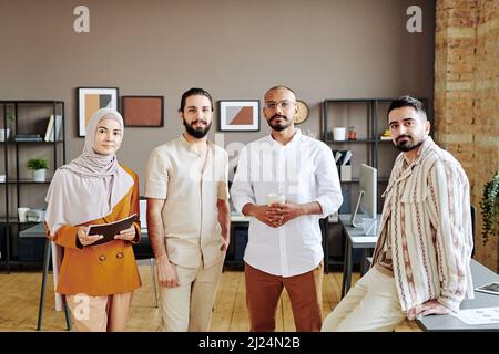 Eine Gruppe junger muslimischer Unternehmer in Casualwear, die vor der Kamera gegen das Innere eines geräumigen, modernen Büros im Freiraum stehen Stockfoto