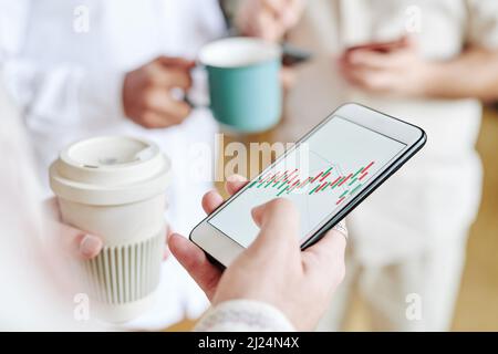 Ein junger, moderner Geschäftsmann mit einer Tasse Kaffee, der die Karte im Smartphone analysiert, während er im Büro gegen Kollegen steht Stockfoto